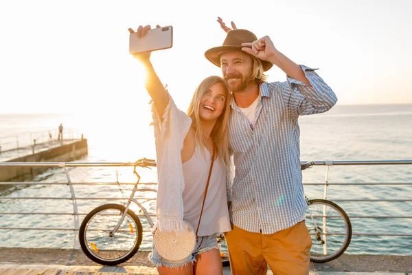 年轻迷人的笑容满面的快乐男人和女人骑自行车旅行 用手机自拍自拍 日落时在海边浪漫的情侣 时髦的时髦装束 朋友们在一起玩乐 — 图库照片