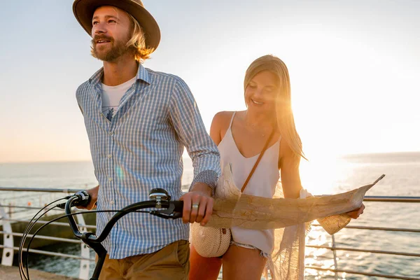 young attractive man and woman traveling on bicycles, holding map, sightseeing, romantic couple on summer vacation by the sea on sunset, boho hipster style outfit, friends having fun together