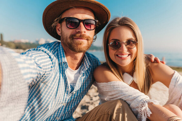 young attractive smiling happy man and woman in sunglasses sitting on sand beach taking selfie photo on phone camera, romantic couple by the sea on sunset, boho hipster style outfit