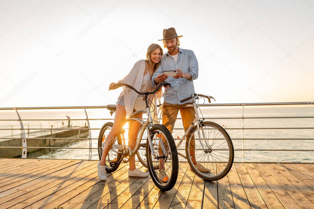 young attractive smiling happy man and woman traveling on bicycles using smartphone, romantic couple by the sea on sunset, boho hipster style outfit, friends having fun together