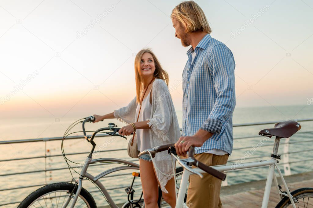 attractive happy couple of friends traveling in summer on bicycles, man and woman with blond hair boho hipster style fashion having fun together, walking by the sea in resort city