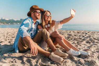 Genç, çekici, güler yüzlü, güneş gözlüklü bir adam ve bir kadın kumsalda oturmuş telefon kamerasında selfie çekiyorlar. Gün batımında deniz kenarında romantik bir çift.