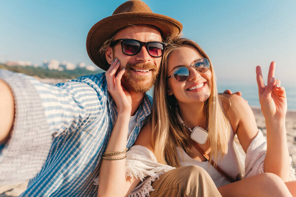 young attractive smiling happy man and woman in sunglasses sitting on sand beach taking selfie photo on phone camera, romantic couple by the sea on sunset, boho hipster style outfit