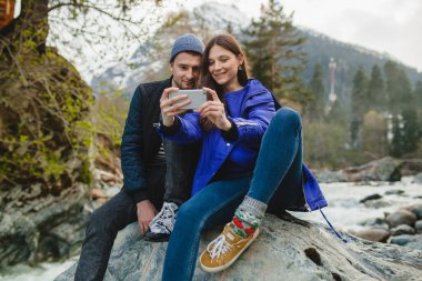 Genç ve güzel hippi çift ellerinde akıllı telefonlar, fotoğraf çekiyorlar, kış ormanlarında nehirde bir kayanın üzerinde oturuyorlar, kot pantolon, kalın palto, spor ayakkabılar, gülümsüyorlar, eğleniyorlar, geziyorlar, yürüyüş yapıyorlar, tatil yapıyorlar.