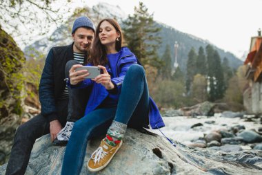 Genç ve güzel hippi çift ellerinde akıllı telefonlar, fotoğraf çekiyorlar, kış ormanlarında nehirde bir kayanın üzerinde oturuyorlar, kot pantolon, kalın palto, spor ayakkabılar, gülümsüyorlar, eğleniyorlar, geziyorlar, yürüyüş yapıyorlar, tatil yapıyorlar.