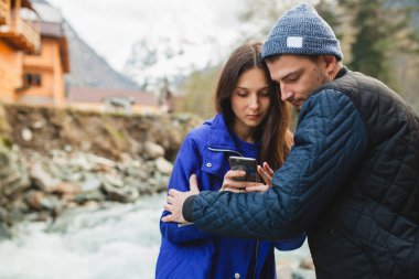 Genç, güzel bir hippi çift ellerinde akıllı telefonlar, fotoğraf çekiyorlar, kış ormanlarında nehirde, sıcak bir palto, gülümsüyor, eğleniyor, seyahat ediyor, yürüyüş yapıyor, tatil yapıyor, örgü örüyor.