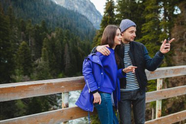 Genç hippi çift kış tatilinde dağlarda, sıcak kıyafetler, örgü şapka, mavi ceket, arka planda ahşap, akıllı telefonu tutuyor ve ona bakıyor, sarılıyor, gülümsüyor, mutlu, seyahat ediyor.