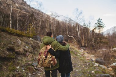 Genç ve güzel hippi erkek ve kadın vahşi doğada birlikte seyahat ediyorlar, ormanda yürüyüş yapıyorlar, sonbahar tarzı kıyafetler, palto, romantizm, tatil, kucaklaşma, arkadan manzara, sırt çantalı