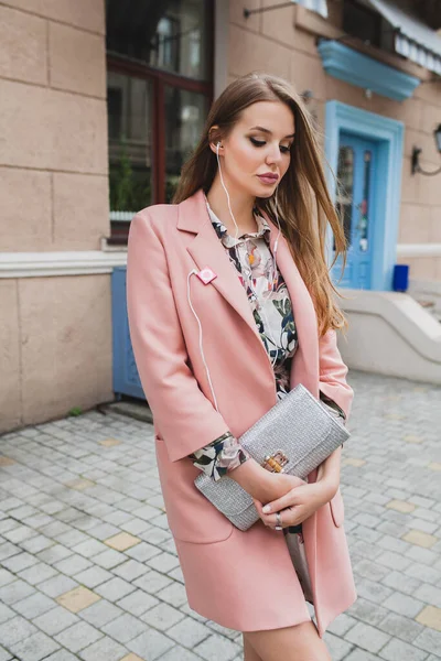 漂亮的年轻貌美的女人穿着粉红的外套走在街上 手里拿着钱包 听着音乐 — 图库照片
