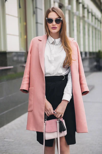 ピンクのコートで街を歩くかなり魅力的なスタイリッシュな笑顔豊かな女性春のファッショントレンド保持財布 エレガントなスタイル サングラスを身に着けている — ストック写真