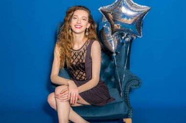 Çok havalı genç hippi güzel bir kadın, gece elbisesi, moda tarzı, mavi arka plan, sandalyede oturan, gümüş balonlar, gülümseyen, mutlu, doğum günü kutlayan