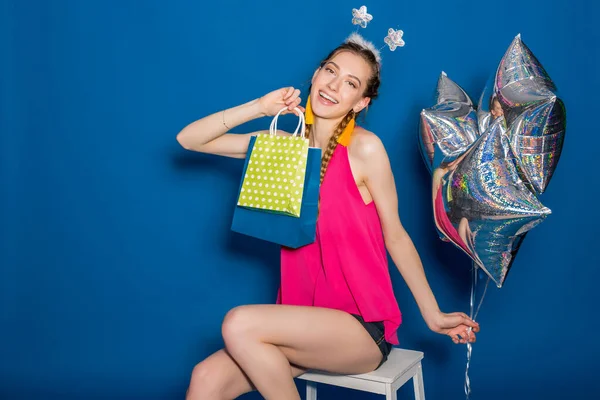 pretty young hipster beautiful woman, colorful outfit, trend fashion style, blue background, holding shopping paper bags, silver star balloons, presents, smiling, happy, celebrating birthday