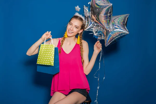 young hipster beautiful woman, colorful outfit, trend fashion style, blue background, holding shopping paper bags, silver star balloons, presents, smiling, happy, celebrating birthday