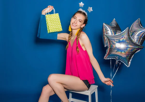 pretty young hipster beautiful woman, colorful outfit, trend fashion style, blue background, holding shopping paper bags, silver star balloons, presents, smiling, happy, celebrating birthday