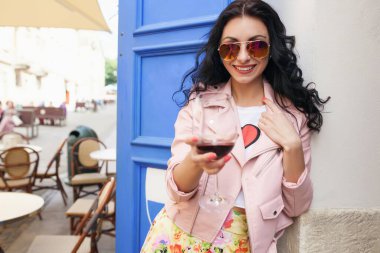 Çekici genç bir kadın yaz tatilinde şarap içiyor. Şehir kafesinde pembe ceketli havalı bir şekilde oturuyor. Gülümsüyor ve güneş gözlüğü takıp eğleniyor.