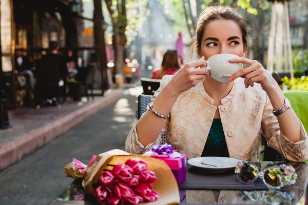 young stylish woman, fashion sunglasses, sitting in cafe, holding drinking cup cappuccino, tulips, happy birthday party, city street, boho outfit, europe vacation, romantic dinner, sunny, smiling