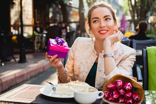 young stylish woman, sitting in cafe, holding present box, smiling, enjoying warm, tulips, happy birthday party, city street, europe vacation, horizontal, glamour outfit, romantic dinner