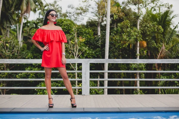 穿着红色夏装的年轻性感女人站在热带酒店的阳台上 棕榈树背景 长长的黑头发 太阳镜 少数民族耳环 太阳镜 向前看 高跟鞋 — 图库照片