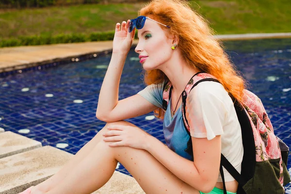 年轻美丽的姜汁女郎坐在游泳池边 背着背包 时髦的衣服 运动鞋 太阳镜 — 图库照片