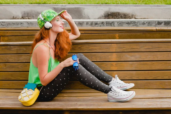 若いスタイリッシュなヒップスタージンジャー女性 ベンチに座って ヘッドフォン 緑のキャップ レギンス スニーカー サングラス 楽しい トレンディーなアパレル ファッショナブルな衣装 — ストック写真