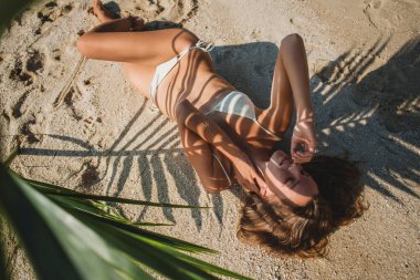 Genç seksi bir kadın kumsalda palmiye yaprağının altında yatıyor, vücudunda sanatsal gölgeler, beyaz bikini mayo, bronzlaşmış ten, ince vücut.