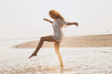 Genç, ince, güzel bir kadın gün batımı kumsalında, neşeli, dans, koşu, bohem kıyafeti, indie tarzı, yaz tatili, güneşli, eğleniyor, pozitif ruh hali, romantik, su sıçratan, siluet, mutlu.
