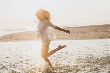 Genç, ince, güzel bir kadın gün batımı kumsalında, neşeli, dans, koşu, bohem kıyafeti, indie tarzı, yaz tatili, güneşli, eğleniyor, pozitif ruh hali, romantik, su sıçratan, siluet, mutlu.