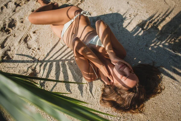 ヤシの木の葉の下の砂のビーチに横たわっている若いセクシーな女性 身体の芸術的な影のパターン 白いビキニスイムスーツ 日焼けした肌 スリムボディ — ストック写真