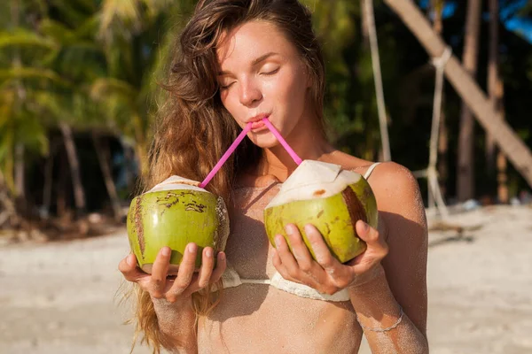 白いビキニ水着姿の若いセクシーな痩せっぽち女性がココナッツを持ち 笑顔で 熱帯ビーチで日光浴をしています スリムなボディ タイでの休暇 自然の美しさ 夏のスタイル 皮膚の砂 — ストック写真