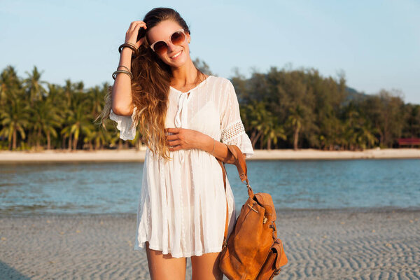 slim beautiful woman in white dress on tropical beach on sunset holding leather backpack. girl happy smiling on vacation, summer style. natural beauty, long hair, boho hippie accessories