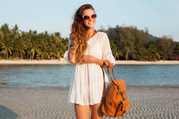 slim beautiful woman in white dress on tropical beach on sunset holding leather backpack. girl happy smiling on vacation, summer style. natural beauty, long hair, boho hippie accessories