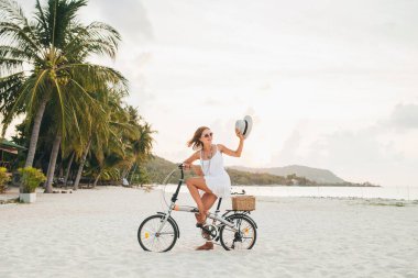 Beyaz elbiseli, tropik plajda bisiklete binen, şapka takan ve güneş gözlüklü genç ve çekici bir kadın yaz tatilinde Tayland 'da, boho moda tarzında seyahat ediyor.