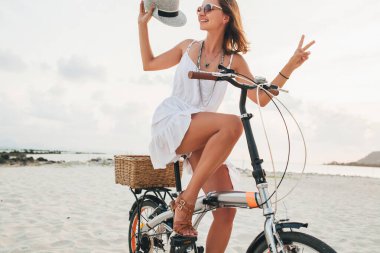 Beyaz elbiseli, tropik plajda bisiklete binen, şapka takan ve güneş gözlüklü genç ve çekici bir kadın yaz tatilinde Tayland 'da, boho moda tarzında seyahat ediyor.