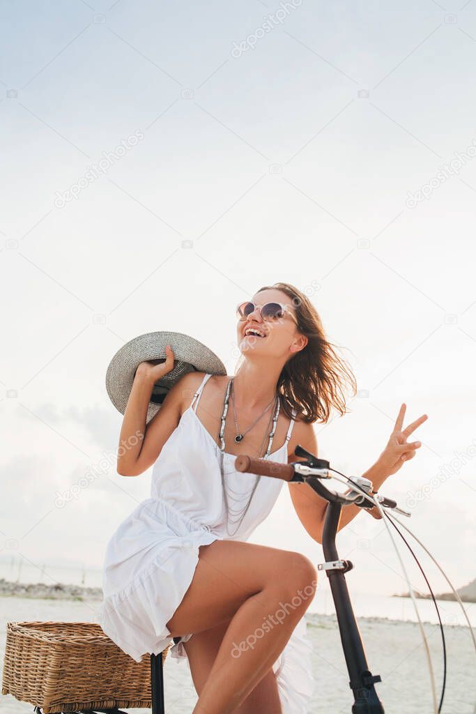 young attractive smiling woman in white dress riding on tropical beach on bicycle wearing hat and sunglasses traveling on summer vacation in Thailand, boho fashion style