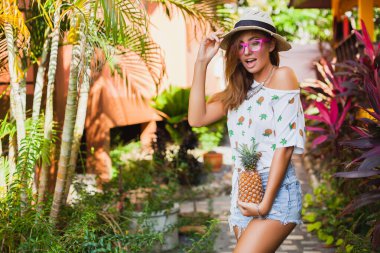 Yazın basılı tişörtle gezen çekici bir kadın hasır şapka takıyor, elleri ananas tutuyor.
