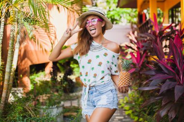 Yazın basılı tişörtle gezen çekici bir kadın hasır şapka takıyor, elleri ananas tutuyor.