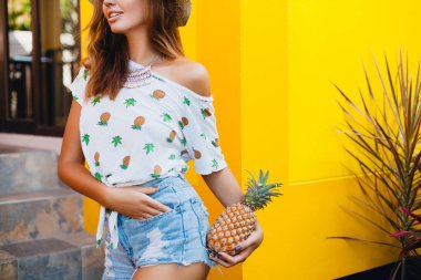 yaz tatilinde çekici bir kadının kot şortu içinde kalçaları ve kalçaları, sıska vücut, ananas, meyve diyeti detoksu, bronzlaşmış ten, sarı arka plan
