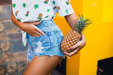 yaz tatilinde çekici bir kadının kot şortu içinde kalçaları ve kalçaları, sıska vücut, ananas, meyve diyeti detoksu, bronzlaşmış ten, sarı arka plan