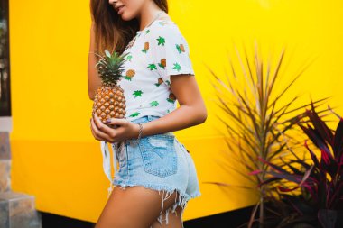 yaz tatilinde çekici bir kadının kot şortu içinde kalçaları ve kalçaları, sıska vücut, ananas, meyve diyeti detoksu, bronzlaşmış ten, sarı arka plan