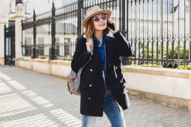 Genç, şık, güzel bir kadın gülümsüyor, mutlu, şaşırmış, telefonuyla konuşuyor koyu mavi ceket ve kot gömlek giyiyor, pembe güneş gözlüğü takıyor, şapka ve sırt çantasıyla, güneşli bir gün, güzel bir hava, şehir caddesi