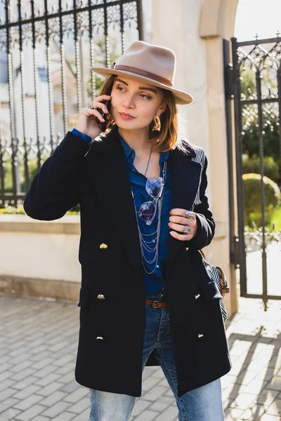 若いスタイリッシュなきれいな女性は笑顔と彼女の携帯電話で話して ダークブルーのコートとジーンズを着て 帽子とバックパックで 晴れた日 天気の良い日 街の通り — ストック写真