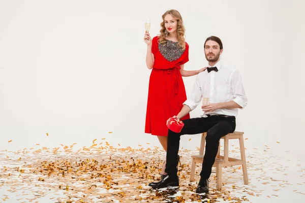 young stylish couple in love on white background holding glasses and drinking champagne, celebrating new year, wearing red dress, fashion jewelry, bow tie, happy disco party, having fun