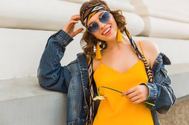şık, çekici bir kadın hippi tarzı kıyafetler giyiyor sarı kot ceket, moda aksesuarlar, güneş gözlükleri, gülümseme, çiçek tutma, sokak modası, bahar moda modası