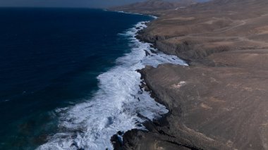  Kuvvetli dalgalarla kayalıklara çarpan kayalık kıyı şeridi berrak bir gökyüzü altında, derin mavi okyanus ve çorak kara arasındaki zıtlığı gözler önüne seriyor..