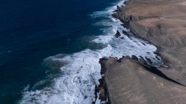  Kuvvetli dalgalarla kayalıklara çarpan kayalık kıyı şeridi berrak bir gökyüzü altında, derin mavi okyanus ve çorak kara arasındaki zıtlığı gözler önüne seriyor..