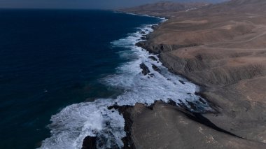  Kuvvetli dalgalarla kayalıklara çarpan kayalık kıyı şeridi berrak bir gökyüzü altında, derin mavi okyanus ve çorak kara arasındaki zıtlığı gözler önüne seriyor..