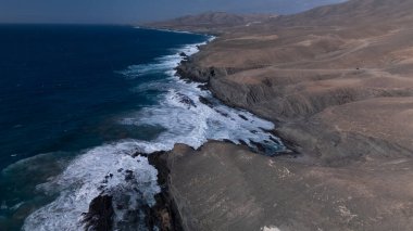  Kuvvetli dalgalarla kayalıklara çarpan kayalık kıyı şeridi berrak bir gökyüzü altında, derin mavi okyanus ve çorak kara arasındaki zıtlığı gözler önüne seriyor..