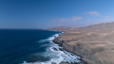  Kuvvetli dalgalarla kayalıklara çarpan kayalık kıyı şeridi berrak bir gökyüzü altında, derin mavi okyanus ve çorak kara arasındaki zıtlığı gözler önüne seriyor..