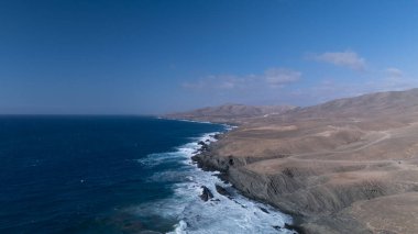  Kuvvetli dalgalarla kayalıklara çarpan kayalık kıyı şeridi berrak bir gökyüzü altında, derin mavi okyanus ve çorak kara arasındaki zıtlığı gözler önüne seriyor..