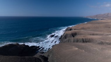  Kuvvetli dalgalarla kayalıklara çarpan kayalık kıyı şeridi berrak bir gökyüzü altında, derin mavi okyanus ve çorak kara arasındaki zıtlığı gözler önüne seriyor..